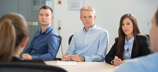 Skupina tří lidí sedí u stolu v kancelářském prostředí. Všichni se dívají směrem k mluvčímu. Jsou oblečeni v košilích, muž vlevo má tmavou košili, dva další mají světlé košile. Na stole je prázdný blok a papíry.