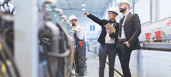 Ein Mann und eine Frau in Schutzhelmen stehen in einer Fabrikhalle. Die Frau zeigt mit dem Finger auf eine Maschine, während sie ein Tablet hält. Ein weiterer Mann in Arbeitskleidung arbeitet im Hintergrund.