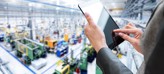 Eine Person hält ein Tablet in der Hand und tippt darauf, im Hintergrund ist eine Fabrikhalle mit Maschinen und Produktionsanlagen unscharf zu sehen.