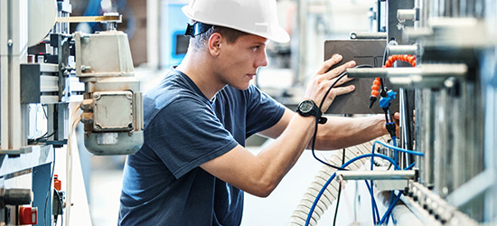 Ein junger Mann in einem weißen Helm arbeitet an einer Maschinensteuerung. Er steht vor einem technischen Gerät und ist konzentriert mit seinen Händen an einer Steuerung beschäftigt. Um ihn herum sind verschiedene Kabel und Apparate zu sehen.