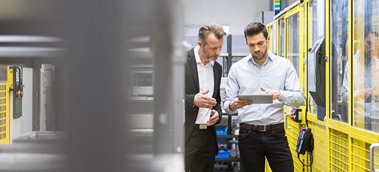 Zwei Männer stehen in einer modernen industriellen Umgebung und sehen auf ein Tablet. Der Mann links trägt einen Anzug, während der Mann rechts ein helles, lässiges Hemd anhat. Im Hintergrund sind gelbe Wände und Maschinen sichtbar.