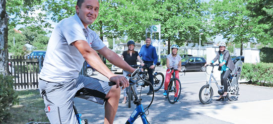 Eine Gruppe von fünf Personen steht und fährt auf Fahrrädern an einem sonnigen Tag. Im Vordergrund ist ein Mann auf einem blauen Fahrrad zu sehen, der in die Kamera lächelt. Im Hintergrund sind drei weitere Personen auf Fahrrädern, während eine Person daneben steht. Bäume und parkende Autos sind im Bild sichtbar.