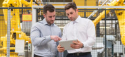 Zwei Männer in Geschäftskleidung stehen in einer Industrieumgebung und betrachten gemeinsam ein Tablet. Im Hintergrund sind gelbe Roboterarme sichtbar.