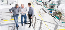 Three men in gray shirts are standing on a staircase, reviewing documents together. They are in a modern industrial setting with machinery visible in the background.
