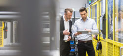 Two men are discussing while looking at a tablet in an industrial setting, surrounded by machinery and yellow safety barriers.