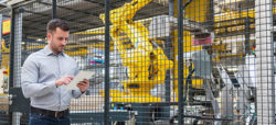 A man stands in front of a safety gate, using a tablet. Behind him, a yellow industrial robotic arm and machinery are visible in a factory setting.