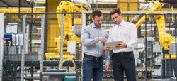 Two men are discussing something on a tablet in a factory setting, surrounded by industrial equipment and robotic arms in the background. One man is wearing a light gray shirt and the other a white shirt, both are engaged in the discussion.