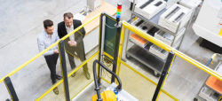 Two men are observing a robotic workstation within an industrial setting, surrounded by safety barriers and equipment. The environment appears to be a manufacturing facility.