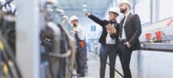 A businesswoman and a businessman are discussing in an industrial setting, pointing at machinery while taking notes on a tablet. Two workers in the background are engaged in tasks related to the equipment. All individuals are wearing safety helmets.