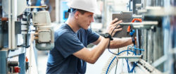A young man wearing a white hard hat is focused on operating machinery in an industrial setting. He is adjusting equipment with visible wires and components around him.