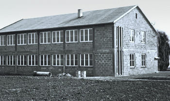 A black and white photograph of a two-story building with a flat roof and multiple windows, situated in a field. The building features a stone exterior and appears to be a simple, utilitarian structure.