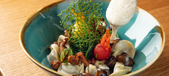 A colorful dish presented in a turquoise bowl features pasta, various mushrooms, a green garnish, a small tomato, and a light foam being drizzled on top.