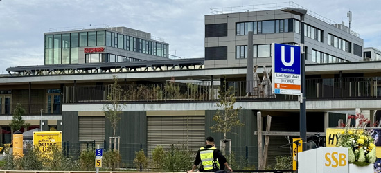 A modern urban setting featuring a city U-Bahn station sign, surrounded by contemporary buildings. There are construction elements visible, with greenery and a figure in a security uniform in the foreground.