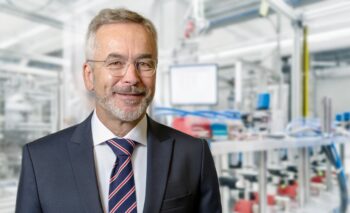 Retrato de un hombre de mediana edad con barba y gafas, vestido con un traje oscuro y una corbata a rayas. Está sonriendo y de fondo se observa un entorno industrial con maquinaria y equipos.