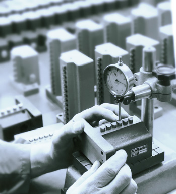 Un operario utiliza un instrumento de medición para verificar la precisión de una pieza de trabajo en un entorno industrial. En el fondo, se pueden ver varias piezas similares dispuestas en una mesa de trabajo.