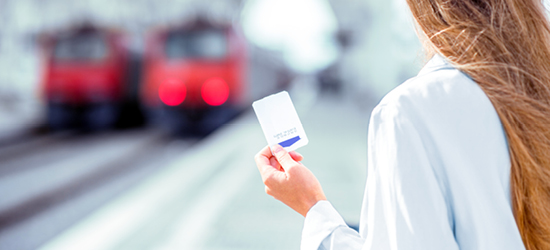 Una persona sostiene una tarjeta en la mano, mirando hacia el tren que se aproxima en una estación. El fondo presenta dos trenes rojos y la escena está iluminada, con un enfoque suave.