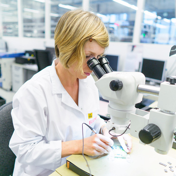 Una mujer con cabello corto y rubio está trabajando con un microscopio en un laboratorio. Lleva una bata blanca y sostiene un objeto en sus manos mientras observa a través del microscopio. En el fondo, se pueden ver computadoras y un ambiente de trabajo limpio y organizado.
