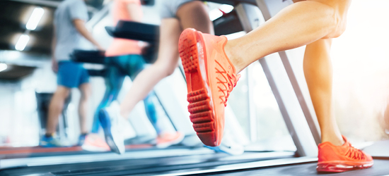 Un homme court sur un tapis de course dans une salle de sport, portant des chaussures de sport rouges. À l'arrière-plan, d'autres personnes s'entraînent également.