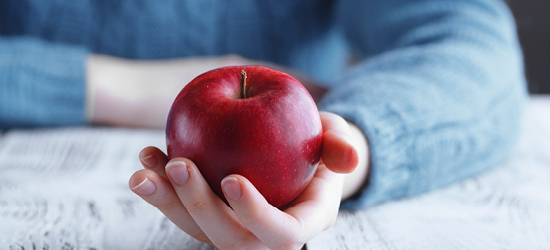 Une main tient une pomme rouge, sur une table en bois. Un pull bleu est visible en arrière-plan.